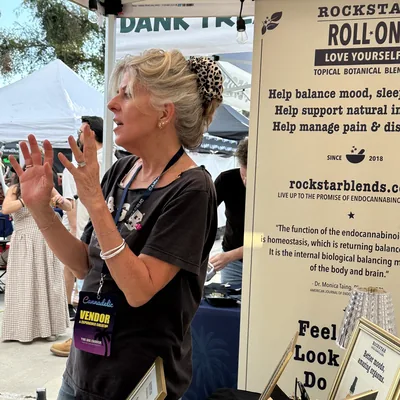 Heather Reilly Hiemstra raises both hands as she talks beside a banner reading Rockstar Roll-On Love Yourself, Since 2018 and rockstarblends.com.