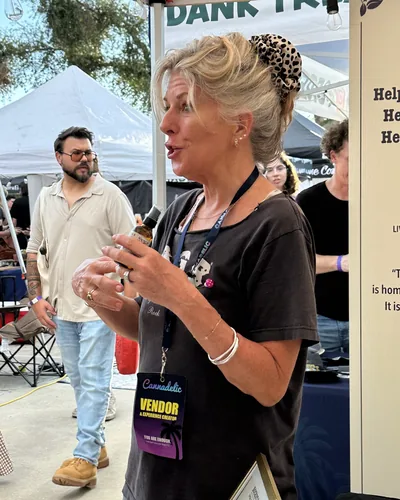 Heather Reilly Hiemstra talks while holding an amber bottle, a Cannadelic vendor badge hanging from her lanyard, with a man and white tents behind her.