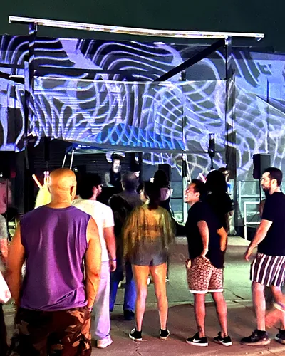 After dark, a small crowd stands before a stage where swirling white and blue light patterns are projected across the canopy and wall.