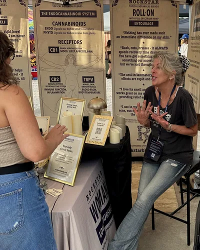 Heather Reilly Hiemstra, seated on a stool, gestures while talking with a visitor across a table of framed signs and candles, under a banner on the endocannabinoid system.