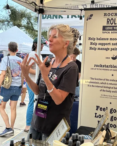 Heather Reilly Hiemstra gestures as she talks, holding a small bottle, with a Cannadelic vendor badge and a banner reading Feel good behind her.