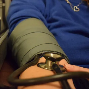 Close photograph of a blood-pressure cuff wrapped around a woman's upper arm in a blue sweater, the chest piece of a stethoscope tucked under its edge.