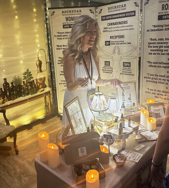 Heather Reilly Hiemstra gestures toward the Rockstar Blends product display—botanical oils, gift boxes, and candles—in front of the Rockstar Woman and endocannabinoid-system banners at Cannadelic Night Market.