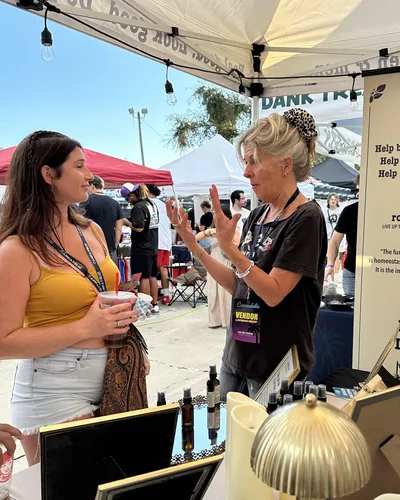 Heather Reilly Hiemstra gestures while talking with a smiling woman holding a drink at the booth table, other vendor tents behind them in daylight.