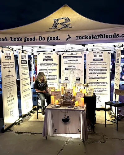 After dark, Heather Reilly Hiemstra sits at her lit tent, which reads Feel good. Look good. Do good. and rockstarblends.com, with banners and candles on the table.