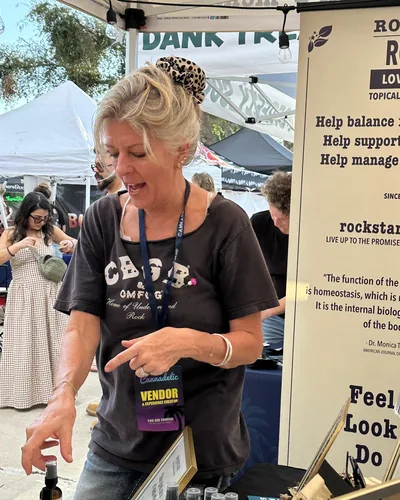 Heather Reilly Hiemstra points toward bottles on her table while talking, wearing a Cannadelic Vendor and Experience Creator badge.