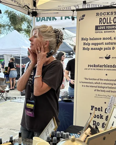 Heather Reilly Hiemstra holds a roller bottle near her jaw in the daylight, wearing a Cannadelic vendor badge, beside a Rockstar Roll-On banner.