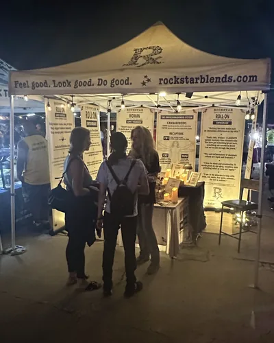 At night, two visitors talk with a woman inside the lit Rockstar Blends tent, banners glowing behind a candlelit table.