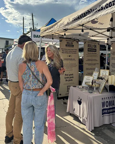 Heather Reilly Hiemstra talks with two visitors under a rockstarblends.com tent in late-afternoon sun, beside banners and a table draped with a Rockstar Woman cloth.