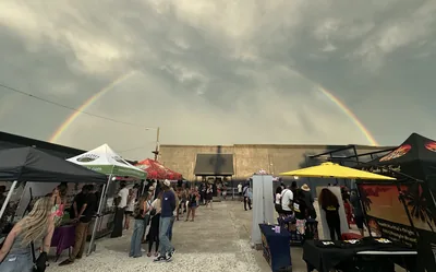 A double rainbow arcs under heavy gray clouds over the block party, with vendor tents and a yellow umbrella lining the street and people walking between them.