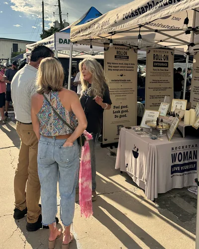 Heather Reilly Hiemstra speaks with two visitors in low golden sun at her tent, with long shadows on the pavement and a table cloth reading Rockstar Woman Love Yourself.